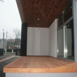 Nowoczesna weranda z drewnianą podłogą i sufitem, duże okna przesuwne, czarna kolumna, widok na ulicę i domy w tle, pochmurny dzień.