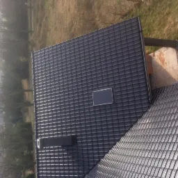 Widok z góry na fragment dachu pokrytego ciemną dachówką ceramiczną z oknem dachowym i kominem wentylacyjnym, sąsiadujący z dachem pokrytym jaśniejszą dachówką, w tle widoczne drzewa i trawa.