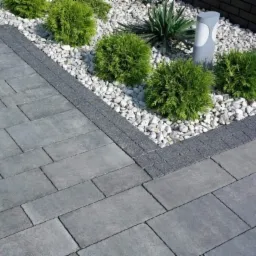 Szara kostka brukowa ułożona w regularny wzór, obrzeże z drobnej kostki, rabata z białymi kamykami, niskimi krzewami i lampą ogrodową.