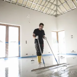 Pracownik w żółtych kaloszach rozprowadza wylewkę samopoziomującą w jasnym, przestronnym pomieszczeniu z oknami zabezpieczonymi folią i widoczną konstrukcją sufitu.