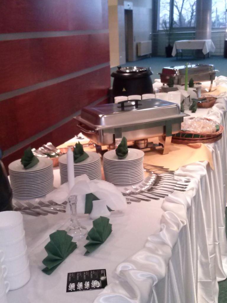 Stół cateringowy z białym obrusem, zastawą, podgrzewaczami, zupą w wazie i dekoracyjnymi serwetkami w kształcie choinek.