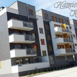 Nowoczesny budynek wielorodzinny z balkonami, wykończony w odcieniach szarości i bieli, z elementami drewnianymi na balkonach i żółtymi okiennicami, sfotografowany w słoneczny dzień.