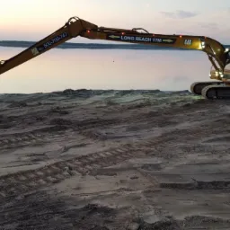 Żółta koparka CAT 320D z długim ramieniem (Long Reach 17M) na piaszczystym terenie, prawdopodobnie nad jeziorem, o zmierzchu. Widoczne ślady gąsienic.