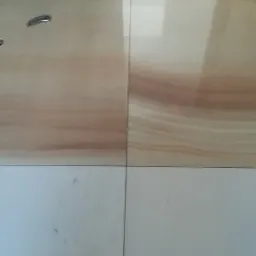 Ułożone płytki podłogowe: górna część w odcieniach beżu z delikatnym wzorem, dolna część biała, widoczne fugi i fragment ściany w trakcie prac wykończeniowych.