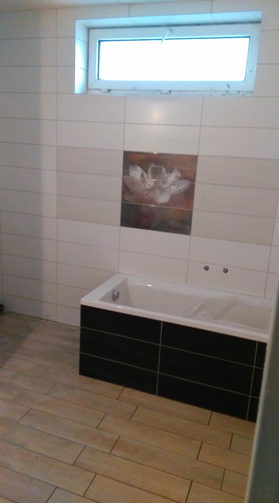 Wykończona łazienka z wanną obudowaną czarnymi płytkami, jasnymi płytkami na ścianach z dekoracyjnym motywem kwiatowym i drewnopodobną podłogą.