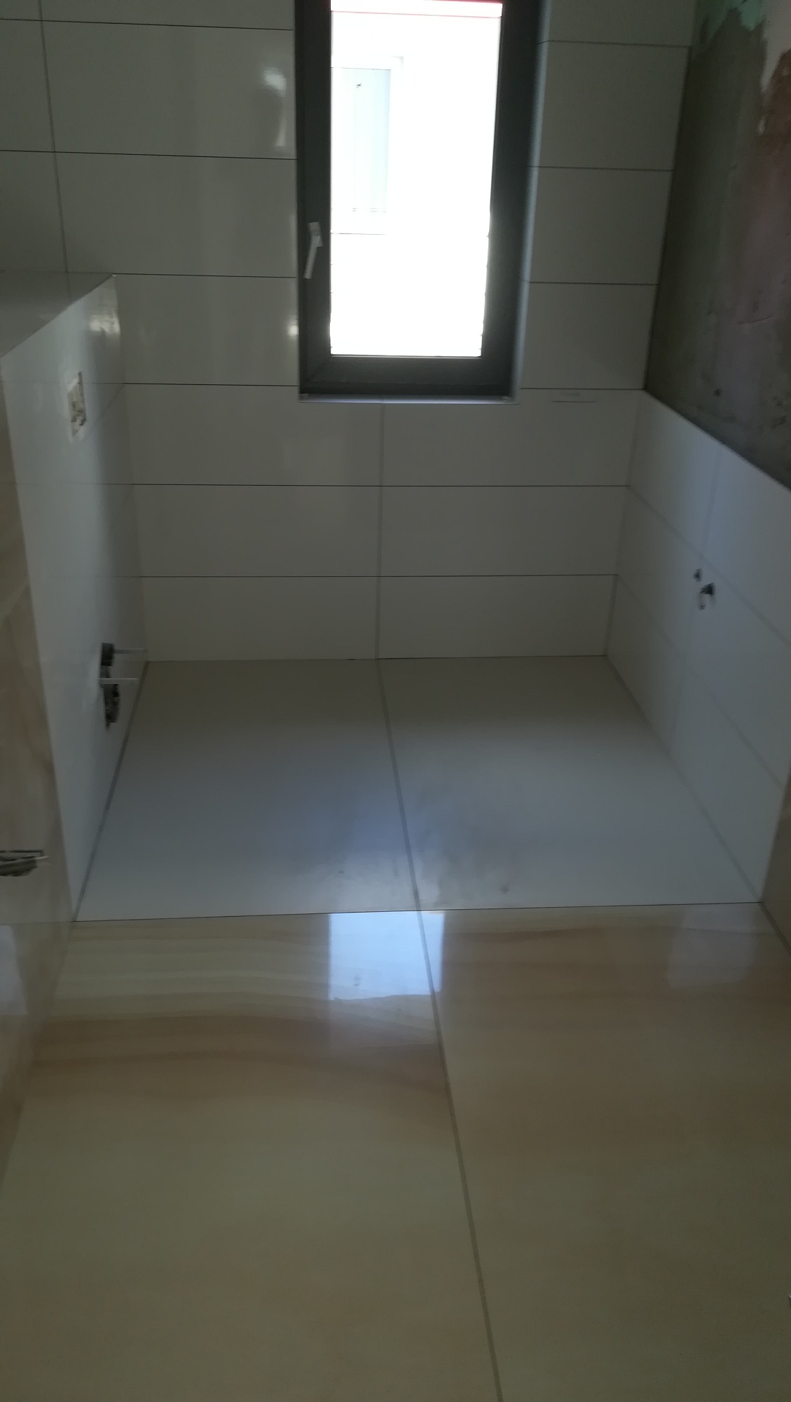 Świeżo wykafelkowana, jasna łazienka z oknem, widoczne przygotowane podejścia pod instalacje, podłoga w odcieniach beżu.