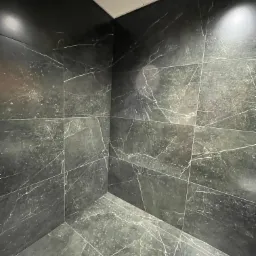 Wykończony narożnik pomieszczenia z ciemnymi płytkami imitującymi marmur z jasnymi żyłami, widoczne fugi i biały sufit kasetonowy.