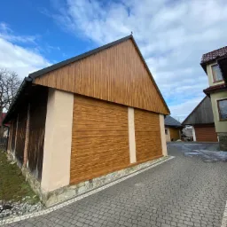 Budynek gospodarczy z drewnianą elewacją w odcieniach brązu, z czarnym dachem i elementami murowanymi, widziany z poziomu kostki brukowej na tle pochmurnego nieba.