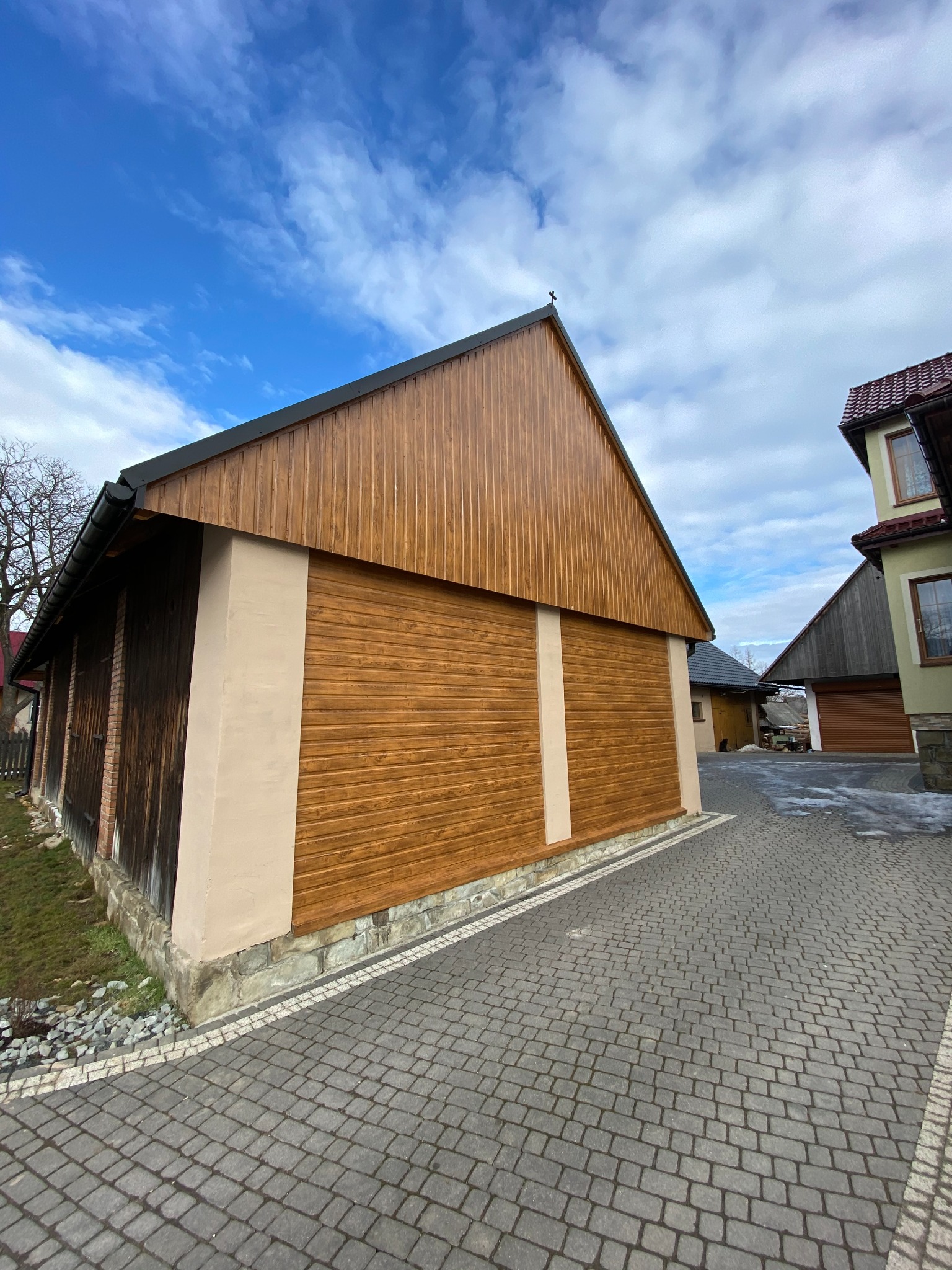 Budynek gospodarczy z drewnianą elewacją w odcieniach brązu, z czarnym dachem i elementami murowanymi, widziany z poziomu kostki brukowej na tle pochmurnego nieba.