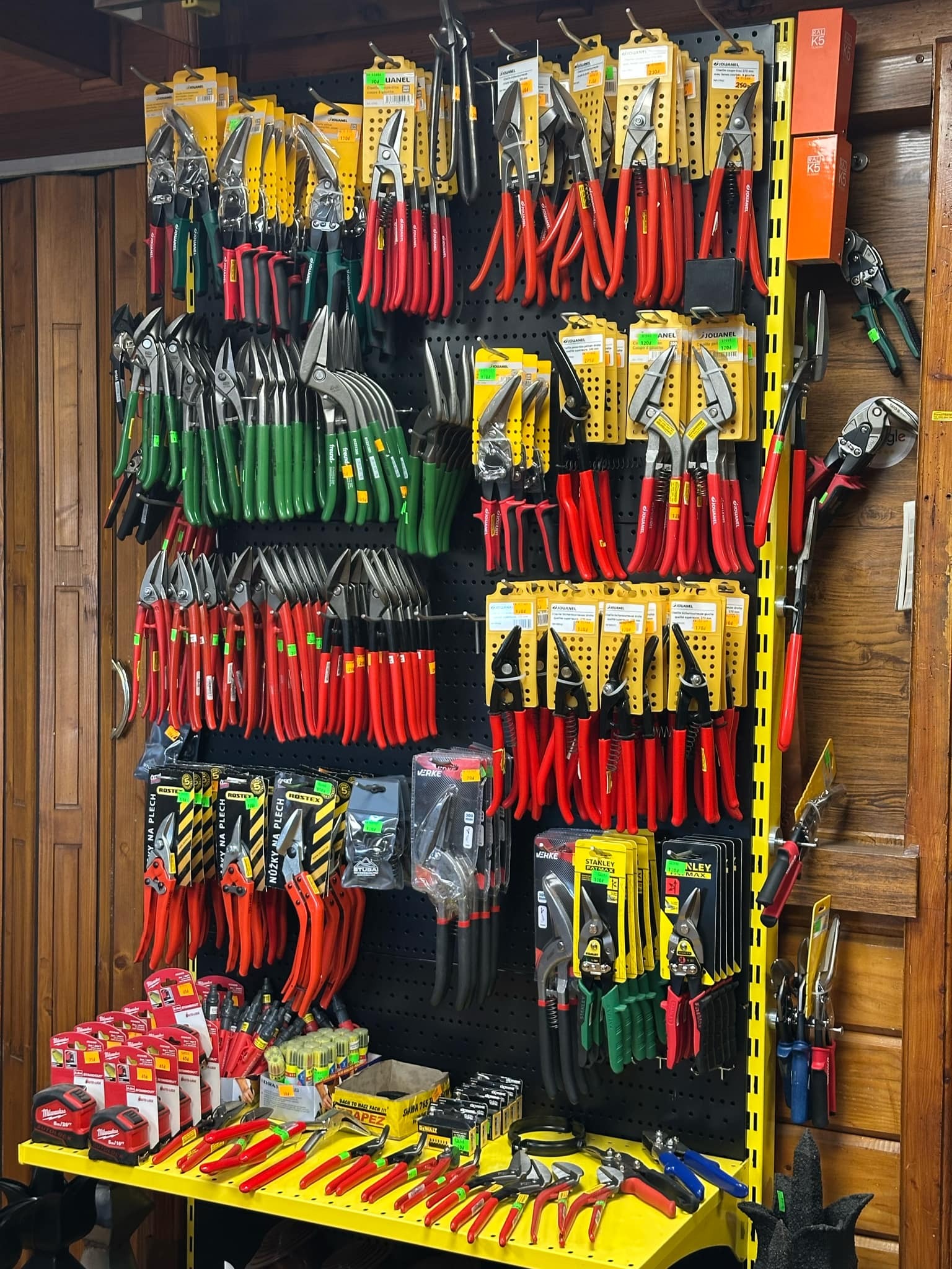 Ekspozycja różnorodnych nożyc do blachy, w tym modeli z czerwonymi, zielonymi i czarnymi rękojeściami, prezentowanych na perforowanej ścianie i żółtym stole w sklepie narzędziowym.