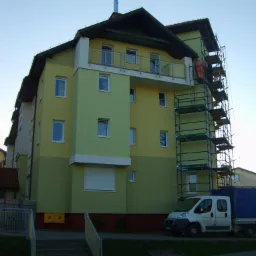 Dwukolorowy budynek wielorodzinny w trakcie prac elewacyjnych, widoczne rusztowanie i pracownik w pomarańczowym kombinezonie, zaparkowany samochód dostawczy w tle.