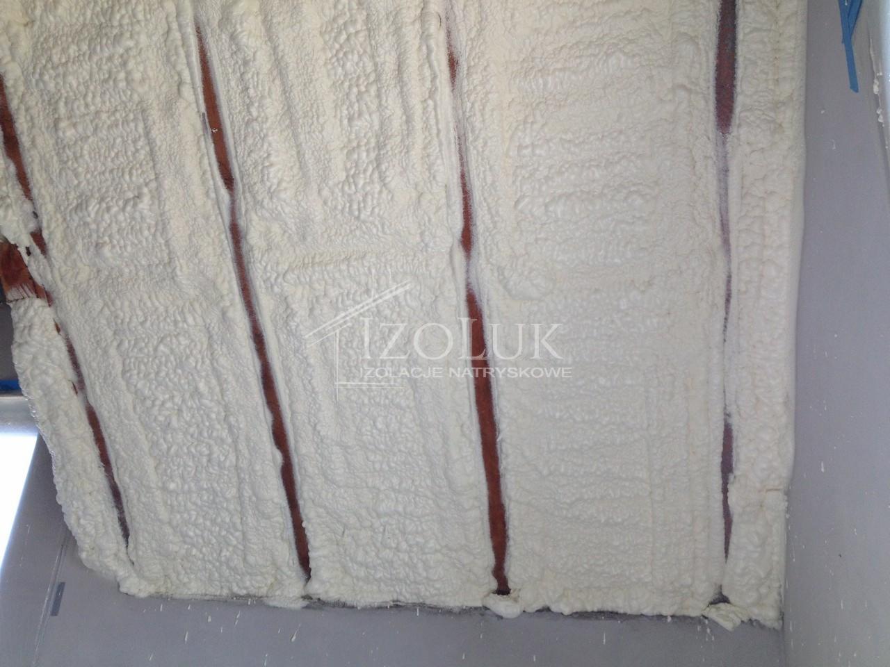 Poddasze z drewnianymi belkami wypełnione białą pianką izolacyjną, widoczne ślady natrysku. Izolacja wykonana w narożniku pomieszczenia.