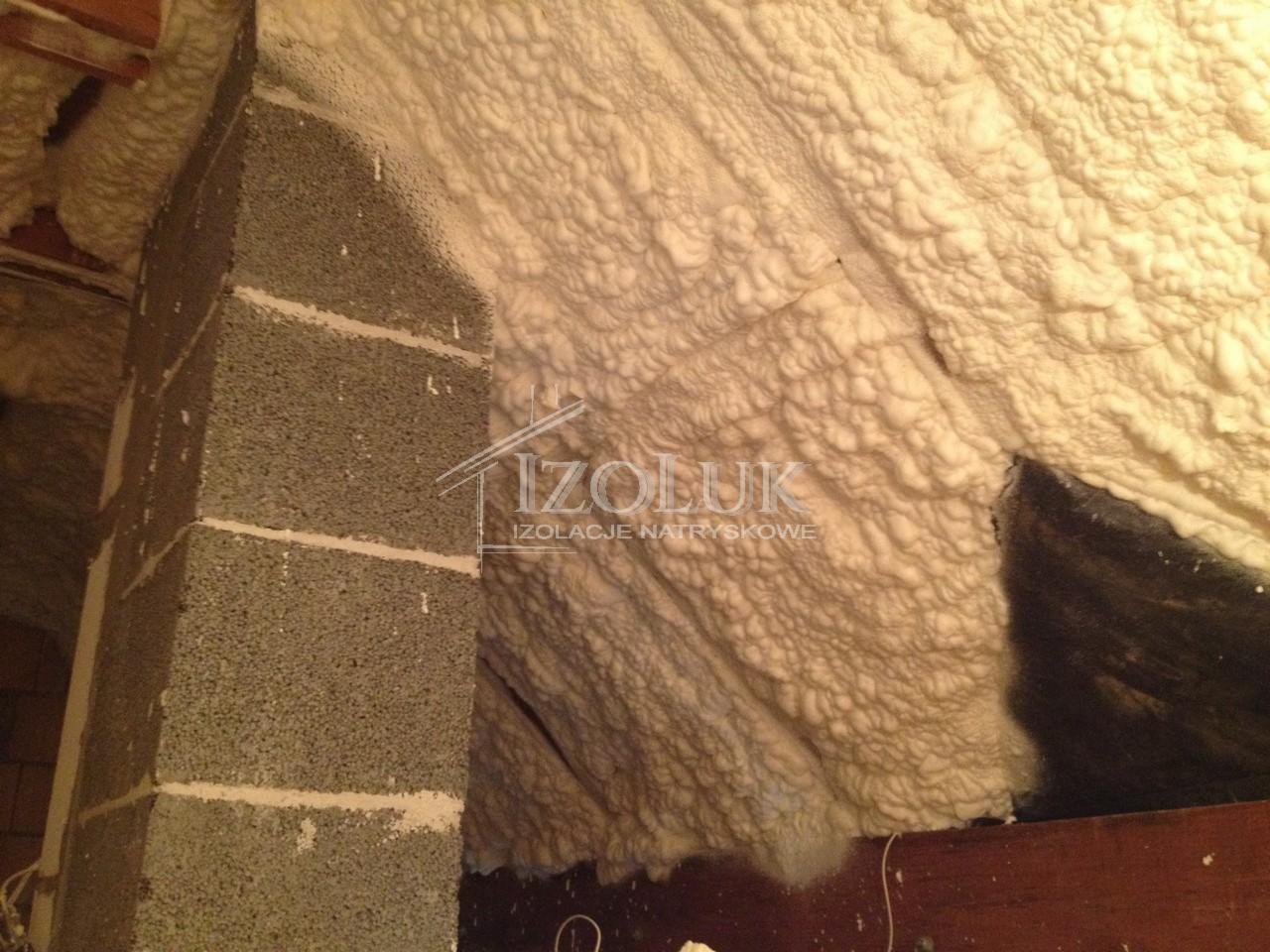 Poddasze po ociepleniu pianką PUR, widoczna struktura piany na skośnych powierzchniach i fragment komina z szarej cegły. Logo firmy IzoLuk.