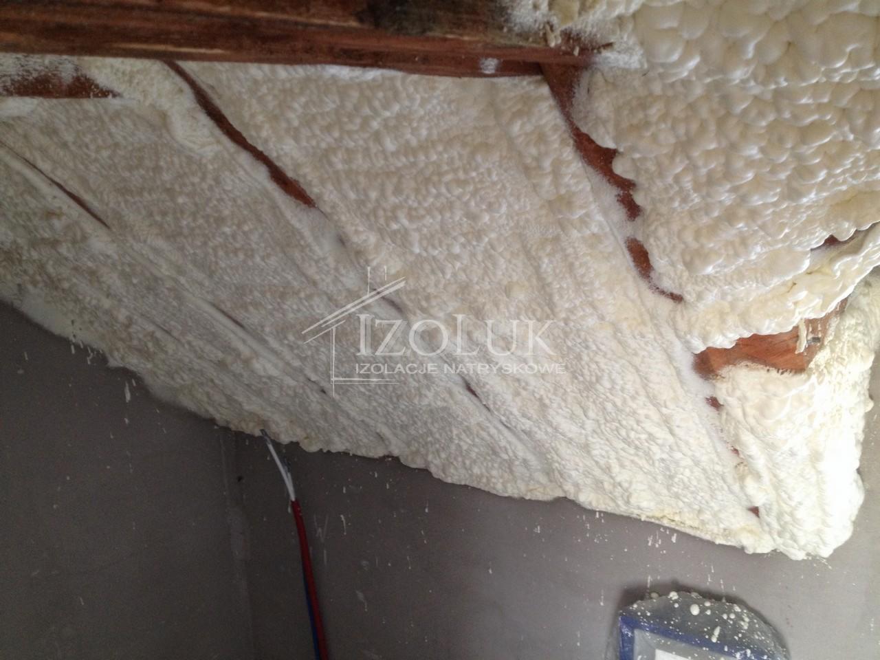 Poddasze ocieplone białą pianką PUR, widoczne drewniane elementy konstrukcji dachu, logo firmy IZOLUK. Izolacja natryskowa wykonana wewnątrz budynku.