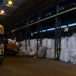 Magazyn z workami wypełnionymi materiałem opałowym, widoczny fragment ciężarówki transportowej i maszyna budowlana w tle.