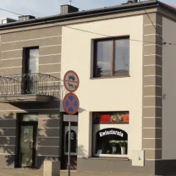 Elewacja dwukondygnacyjnego budynku z balkonem i oknami, z widocznymi szyldami firm: telefony komórkowe i kwiaciarnia, na tle ulicy i znaków drogowych.