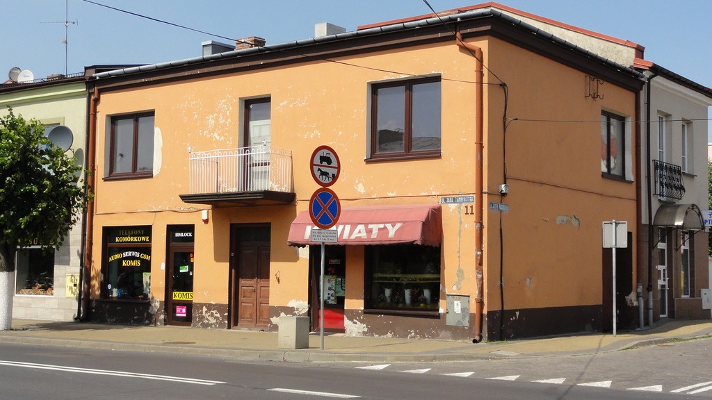 Dwupiętrowy budynek w kolorze pomarańczowym z brązowymi oknami, balkonem z białą balustradą i czerwonym daszkiem nad wejściem do sklepu 'KWIATY', widoczne znaki drogowe.