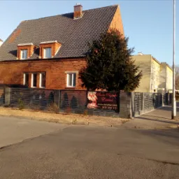 Dom z czerwonej cegły za szarym, poziomym ogrodzeniem panelowym, na którym wisi baner reklamowy salonu kosmetycznego.