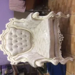 Biały, bogato zdobiony fotel w stylu Ludwika, z tapicerowanym siedziskiem i oparciem, zabezpieczony folią ochronną, widoczny na tle ściany i podłogi.