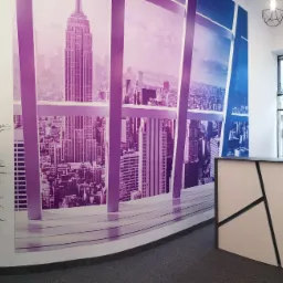 Recepcja w biurze finansowym z fototapetą przedstawiającą panoramę miasta z drapaczami chmur w odcieniach fioletu i błękitu, w tle widoczne plakaty reklamowe z ofertą finansową.
