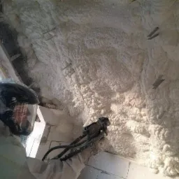 Pracownik w kombinezonie aplikuje pianę PUR na sufit z widocznymi metalowymi elementami montażowymi. Struktura piany przypomina gęstą, białą chmurę.