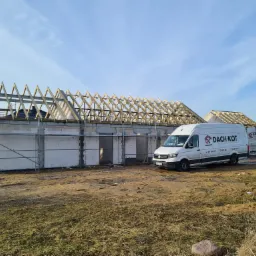 Wznoszona konstrukcja budynku z widoczną drewnianą więźbą dachową, obok zaparkowany biały bus z logo firmy dekarskiej.