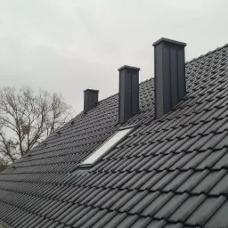 Nowy, ciemnoszary dach z dachówki ceramicznej z trzema kominami wentylacyjnymi i oknem dachowym, widoczne drzewo bez liści na tle pochmurnego nieba.