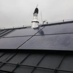 Cztery panele fotowoltaiczne zamontowane na grafitowym, blaszanym dachu z kominem w tle, przy pochmurnym niebie. Widoczne zabezpieczenia przeciwśniegowe.