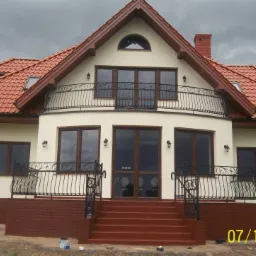 Dwupiętrowy dom z balkonem i czerwonym dachem po renowacji elewacji, z widocznymi schodami wejściowymi i datą wykonania prac na pierwszym planie.