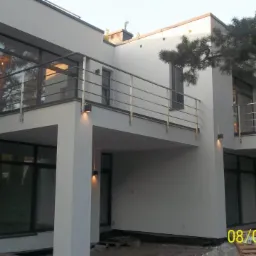 Nowoczesny, dwupiętrowy dom z dużymi oknami i balkonami z chromowanymi balustradami. Elewacja w jasnym odcieniu szarości, oświetlenie zewnętrzne. Widoczne drzewa w tle.