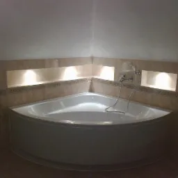 Narożna, biała wanna z baterią i słuchawką prysznicową, otoczona beżowymi płytkami. Nad wanną wnęki z oświetleniem punktowym. Całość w wykończonej łazience.