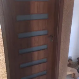 Zamknięte, brązowe drzwi wewnętrzne z pięcioma poziomymi, matowymi przeszkleniami, zamontowane w pomieszczeniu z korkową tapetą i jasną podłogą wyłożoną płytkami.