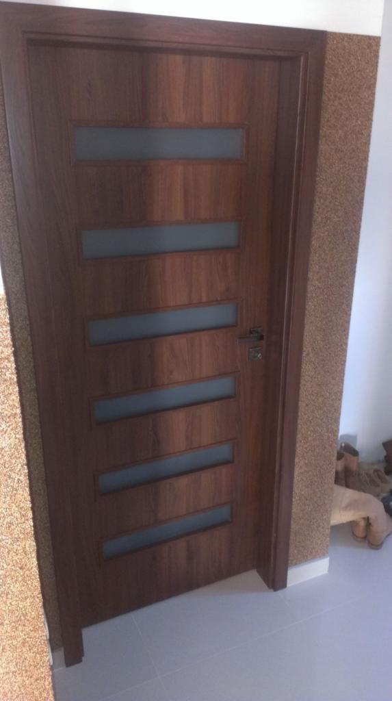 Zamknięte, brązowe drzwi wewnętrzne z pięcioma poziomymi, matowymi przeszkleniami, zamontowane w pomieszczeniu z korkową tapetą i jasną podłogą wyłożoną płytkami.