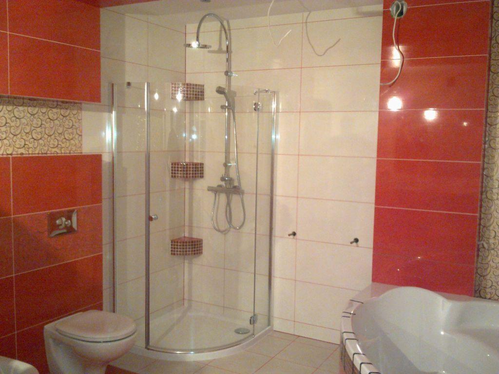 Ukończona łazienka z kabiną prysznicową narożną ze szklanymi drzwiami, czerwono-białe płytki na ścianach z dekoracyjnym pasem, toaleta i wanna z hydromasażem w rogu, kable wystające z sufitu.