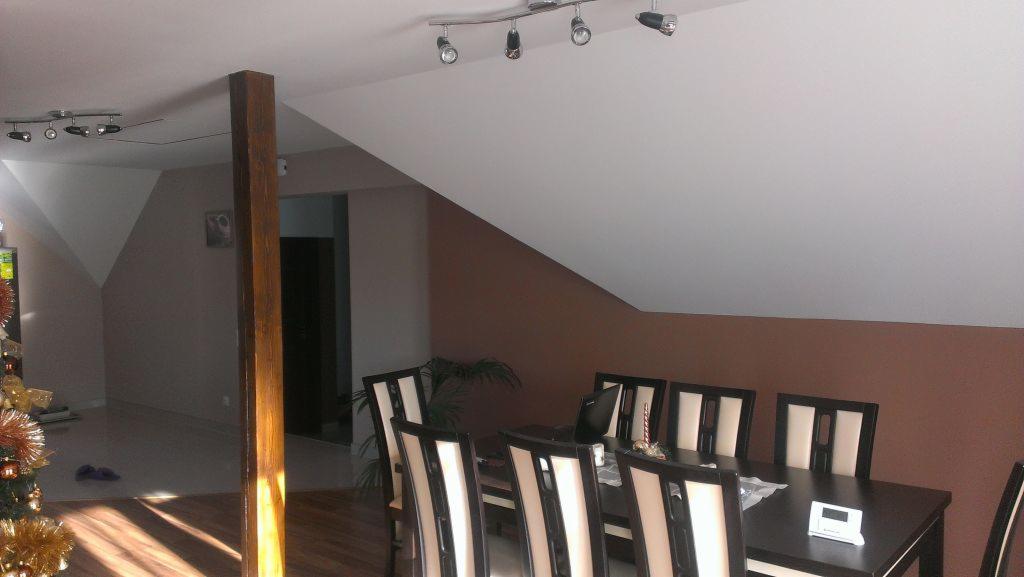 Zabudowa z płyty gipsowo-kartonowej w pokoju na poddaszu, widoczny fragment drewnianego słupa konstrukcyjnego, stół z krzesłami i oświetlenie sufitowe.