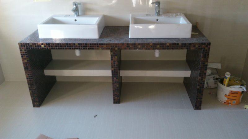 Dwie białe, prostokątne umywalki nablatowe z chromowanymi bateriami, osadzone na blacie wykończonym mozaiką w odcieniach brązu i czerni, z otwartymi półkami poniżej, widok z przodu, ściany...