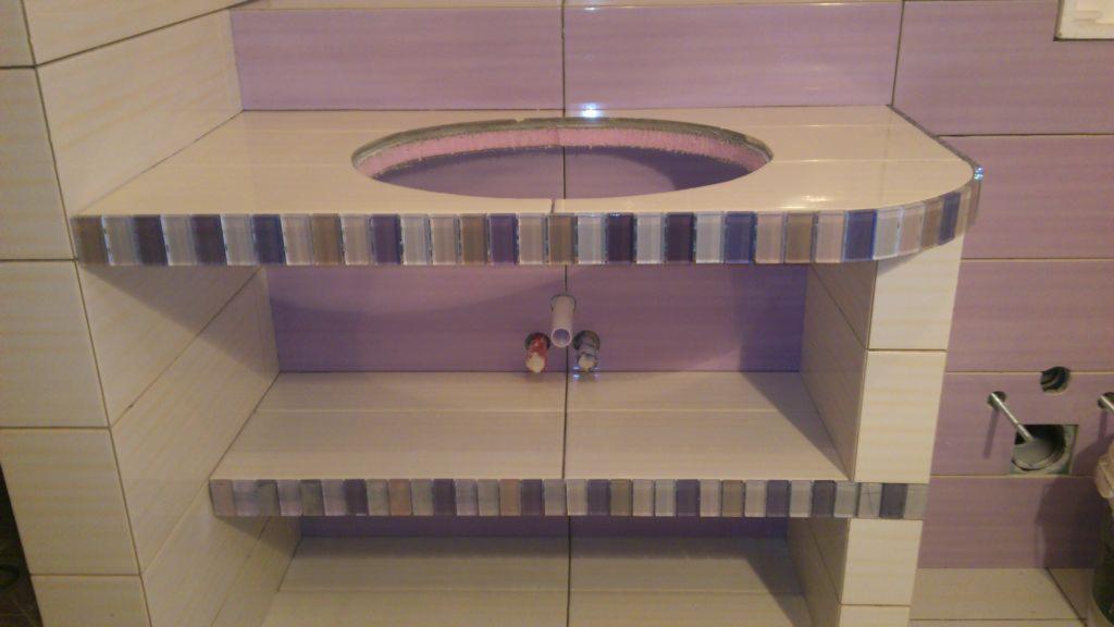 Niedokończona zabudowa umywalki w łazience, wykończona mozaiką w odcieniach fioletu i beżu, widoczne przyłącza wodne i otwór na umywalkę.