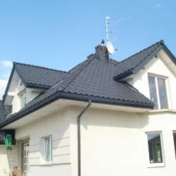 Nowoczesny dom jednorodzinny z ciemną dachówką, antena satelitarna na kominie i pracownik na drabinie. Widok z dołu na elewację i fragment dachu.