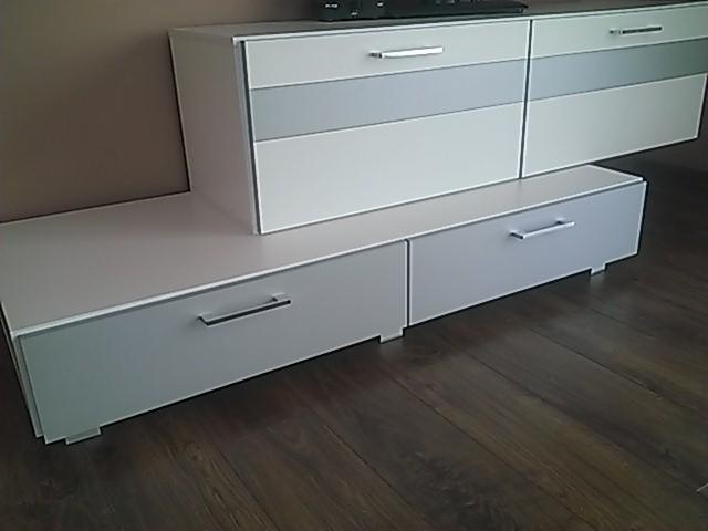 Nowoczesna szafka RTV LUNNA w kolorze białym i szarym, z dwiema szufladami na dole i trzema szufladami u góry, widoczna na tle brązowej ściany i drewnianej podłogi.