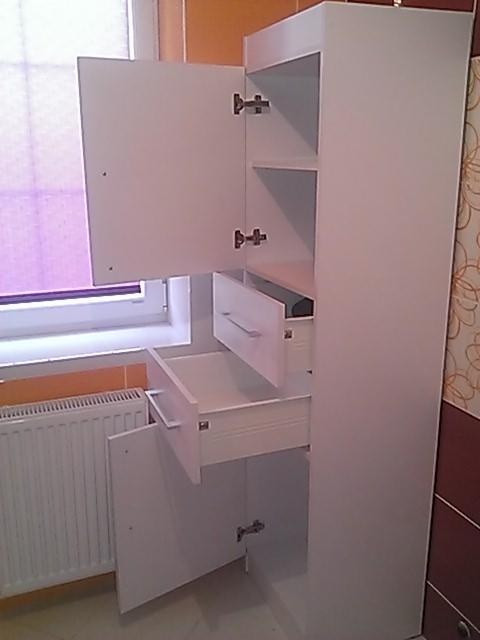 Biała szafka łazienkowa z otwartymi drzwiczkami i szufladami, widoczne zawiasy i wnętrze szafki, grzejnik i okno w tle.