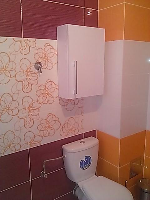 Biała szafka łazienkowa z długim, srebrnym uchwytem, zamontowana nad toaletą w łazience z bordowo-pomarańczowymi płytkami i wzorem kwiatowym.