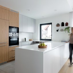 HOME PLAN PROJEKTOWANIE WNĘTRZ JOANNA MIELCZAREK - Minimalistyczna kuchnia z wyspą, fronty szafek w kolorze jasnego drewna i bieli, na wyspie drewniana misa z zielonymi gruszkami, na ścianie półki z dekoracjami etnicznymi i rośliną.