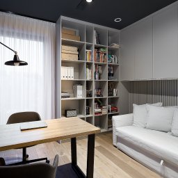 HOME PLAN PROJEKTOWANIE WNĘTRZ JOANNA MIELCZAREK - Nowoczesny gabinet z drewnianym biurkiem, szarymi krzesłami, regałem z książkami i białym wypoczynkiem, oświetlony reflektorami na czarnym suficie.