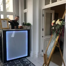 Barman w czarnej koszuli i muszce nalewa napój z butelki do shakera na podświetlanym barze podczas eventu w jasnym pomieszczeniu z dekoracjami kwiatowymi.