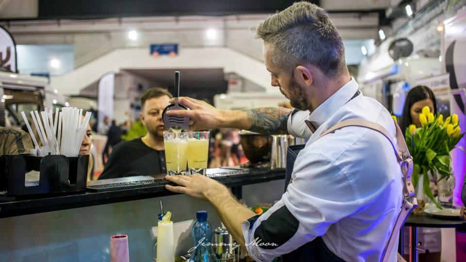 Barmam z tatuażem nalewa drinki do szklanek za barem, w tle widoczni klienci i dekoracje kwiatowe.