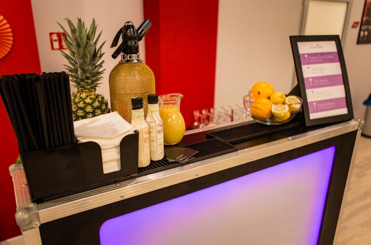 Mobilny bar z podświetleniem LED, ananasem, syfonem w złotej siatce, owocami cytrusowymi w szklanej misie oraz menu drinków.