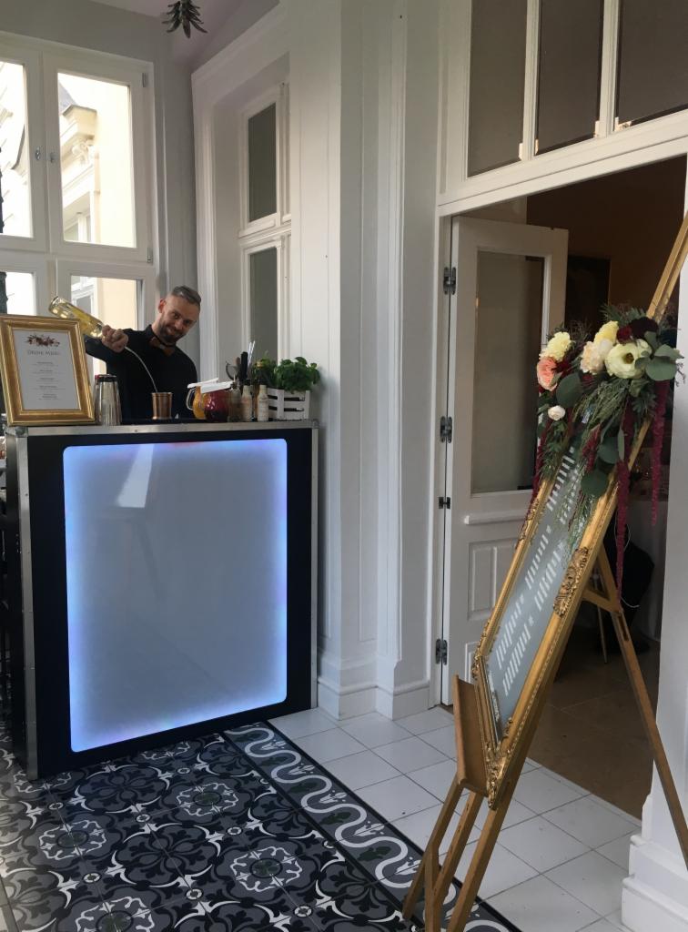 Barman w czarnej koszuli i muszce nalewa napój z butelki do shakera na podświetlanym barze podczas eventu w jasnym pomieszczeniu z dekoracjami kwiatowymi.