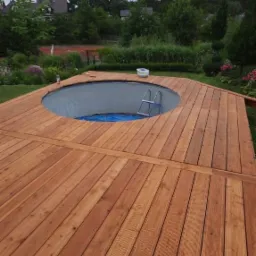 Drewniany taras otaczający okrągły basen z drabinką, widok na ogród z domami w tle.