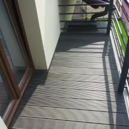 Szary taras kompozytowy na balkonie z metalową balustradą, widok z góry, z widocznym oknem balkonowym z brązową ramą.