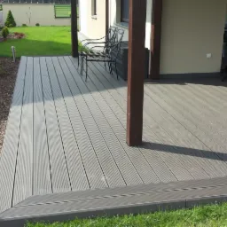 Szary taras kompozytowy z widocznymi ryflowaniami, podparty na drewnianych słupach, z czarnymi metalowymi krzesłami i widokiem na trawnik oraz krzewy, z kotem siedzącym przy drzwiach tarasowych.